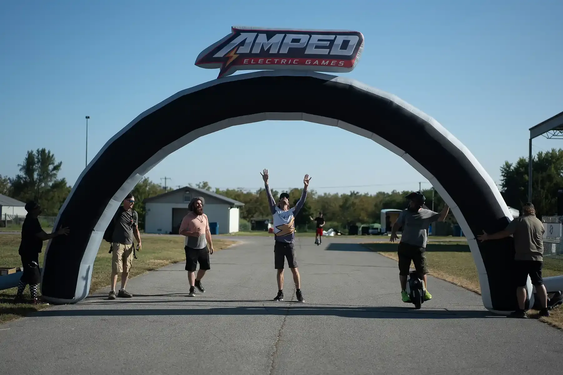 Riders on electric unicycles roll through an inflatable Amped Electric Games arch, one with both arms raised