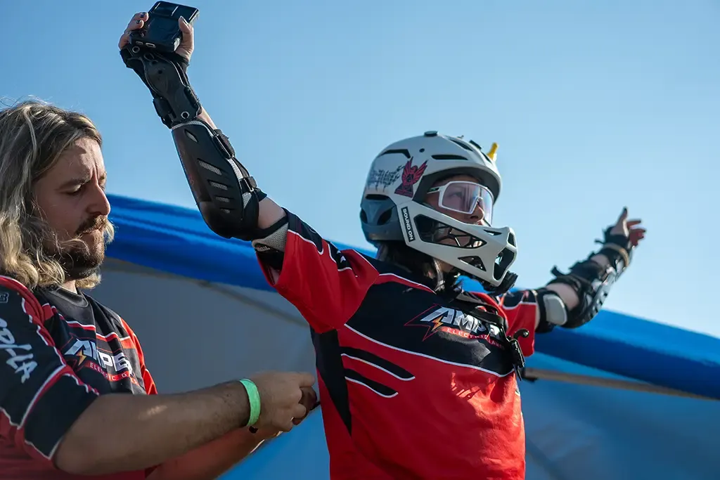 A rider in a helmet and goggles throws both arms up in celebration while a teammate congratulates him