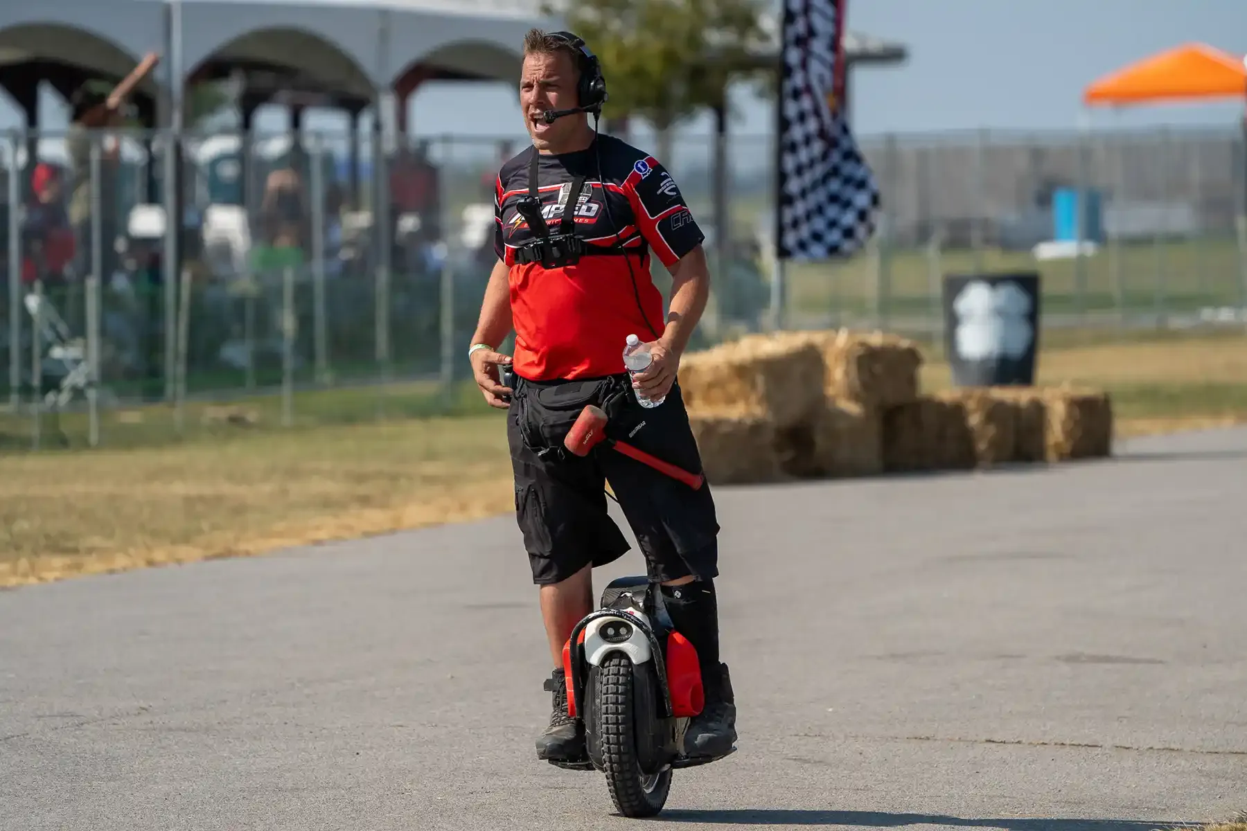 A smiling rider wearing a headset mic rides an electric unicycle past hay bales at a race event