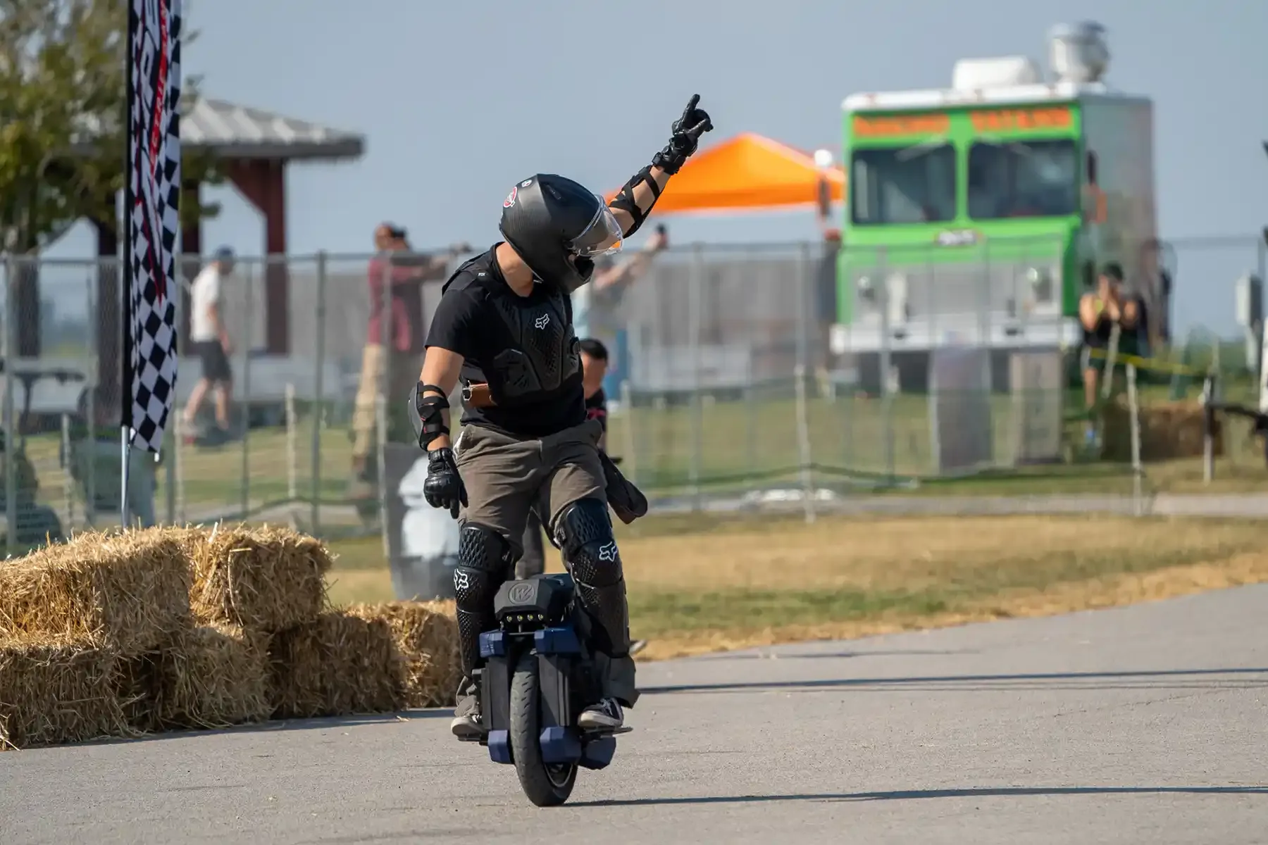 A rider in black gear raises one arm while riding an electric unicycle past hay bales and a food truck
