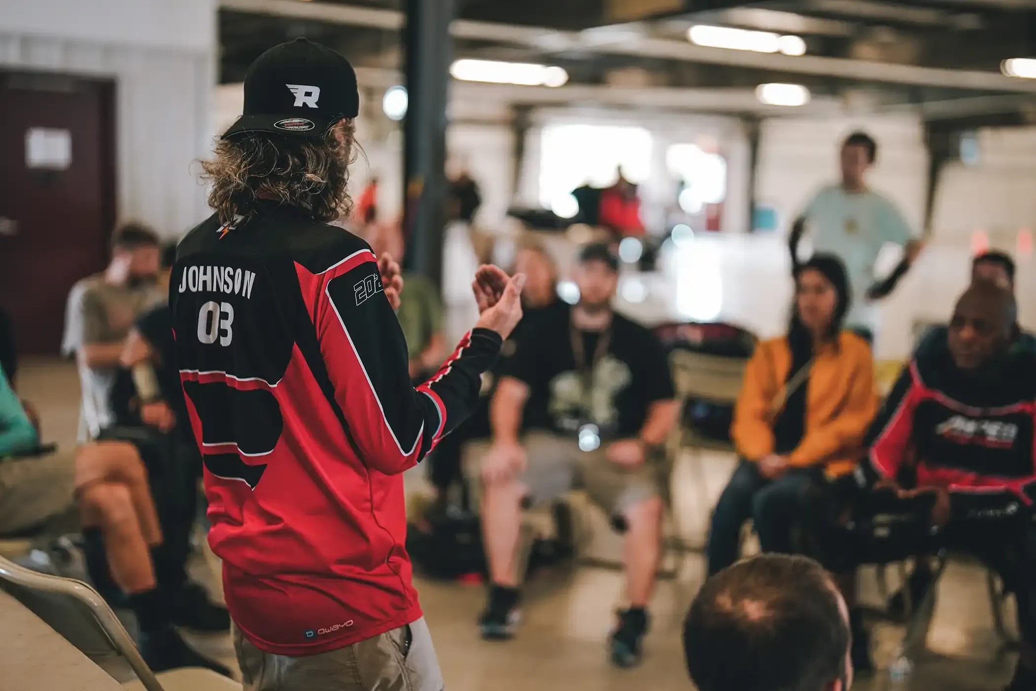 An instructor in a red and black racing jersey numbered 03 speaks to a seated group of riders in a large hall